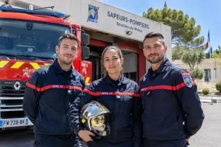 Trois sapeurs-pompiers en uniforme devant une caserne du Sud de la France par une belle journée ensoleillée, avec un camion de pompier rouge et des arbres en arrière-plan.